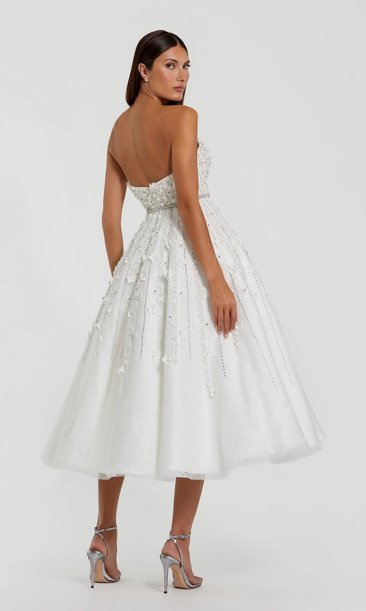 Woman wearing the Mac Duggal 11865 Strapless Champagne Party Dress with embellishments and silver heels, standing on a light background and glancing over her shoulder.
